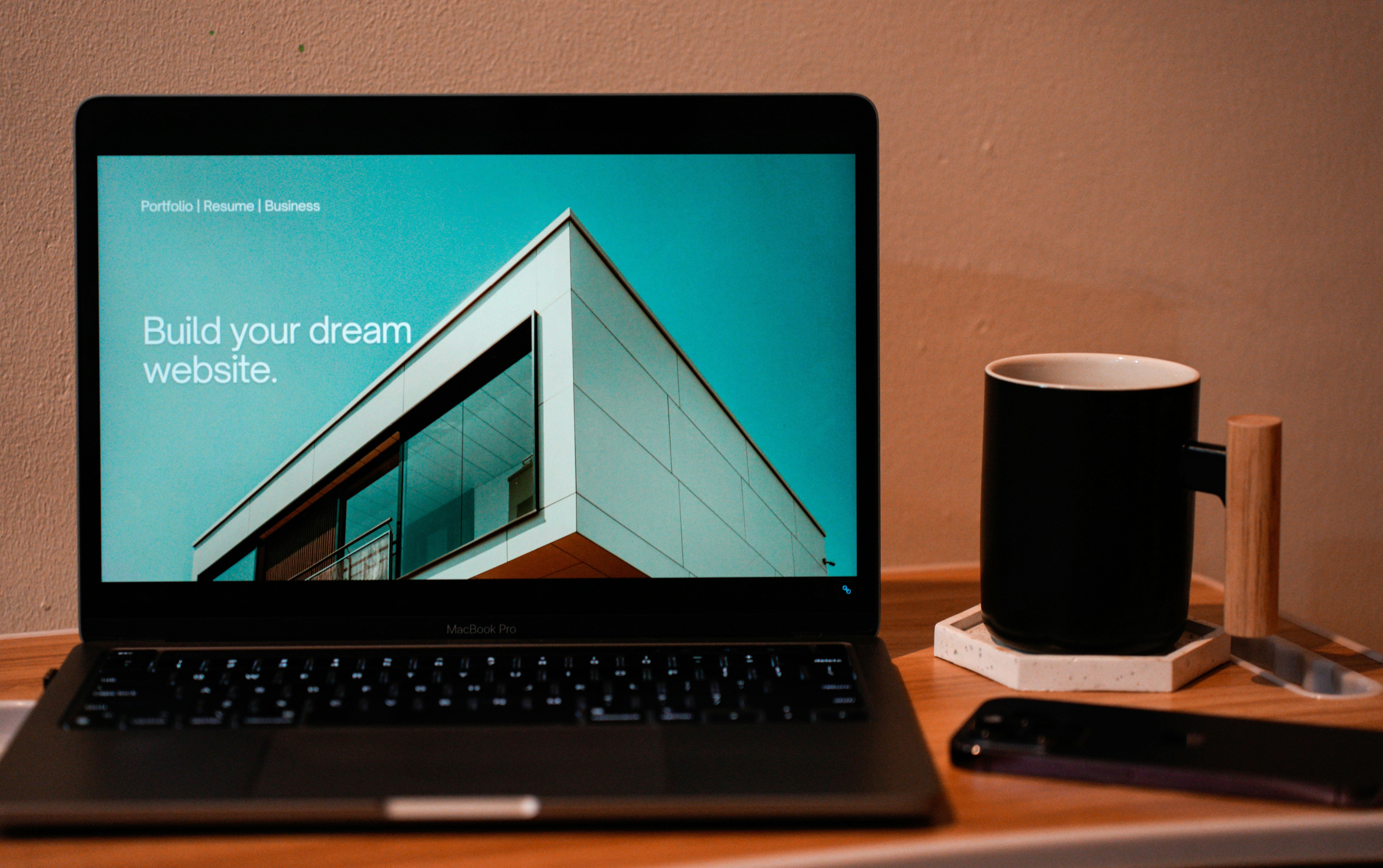 Laptop on wood table displaying a modern building and text 'Build your dream website' with a mug and phone nearby.